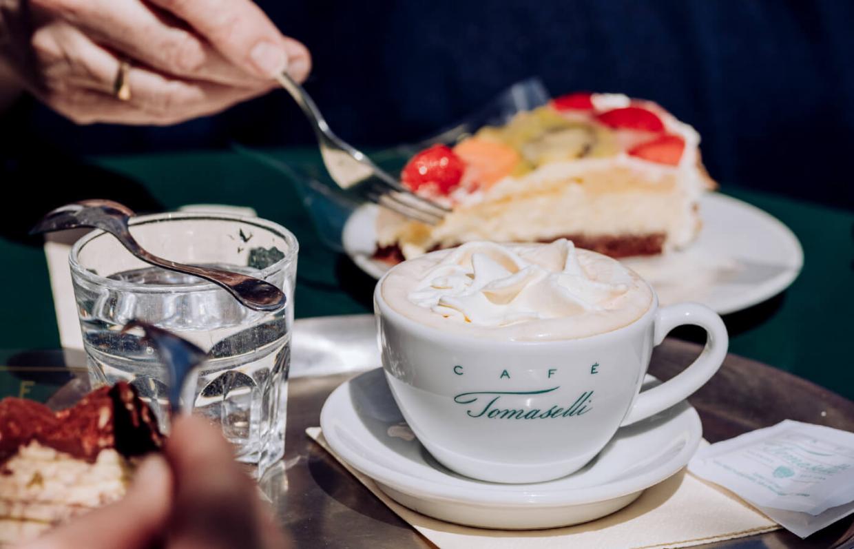 Ein Café Tomaselli Tasse gefüllt mit einem cremigen Getränk, daneben ein Stück Obsttorte auf einem Teller, auf einem Tisch im Freien. Eine Hand mit einem goldenen Ring greift nach einem Löffel, um ein Stück der Torte zu nehmen.