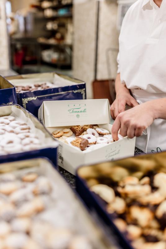 Eine Angestellte im Café Tomaselli packt sorgfältig verschiedene Sorten von frischen Keksen in eine markierte Schachtel, umgeben von weiteren Schachteln mit Gebäck in einer gut ausgestatteten Küche.